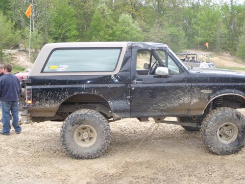 1996 Ford Bronco - A muddy 1996 Ford Bronco with a tan top is seen off-road, with another vehicle in the background.