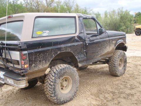 1996 Ford Bronco - This lifted 1996 Ford Bronco, seen in a muddy, off-road setting, features a black body with a tan removable top and large off-road tires.