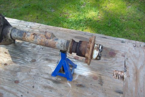 1995 Ford Bronco - A heavily rusted axle housing is supported by two blue jack stands on a weathered wooden surface.