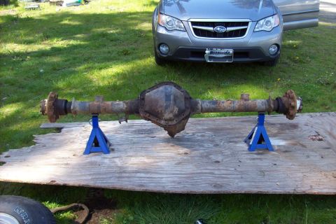 1995 Ford Bronco - A rusty rear axle assembly is displayed on jack stands in a grassy yard, with a Subaru Outback in the background.