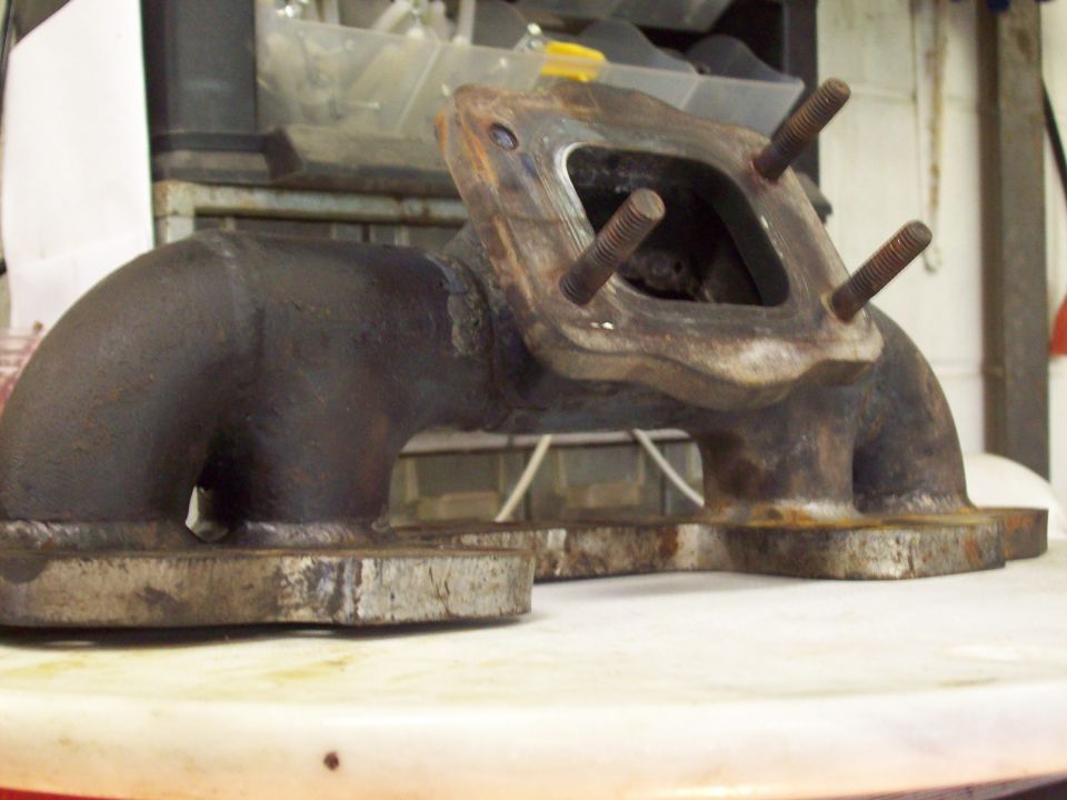 Rusty cast-iron exhaust manifold with four studs in the flange on a shop stool