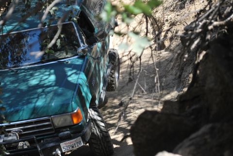 1996 Ford Bronco - Green SUV navigating a rough dirt trail with trees and rocks around.