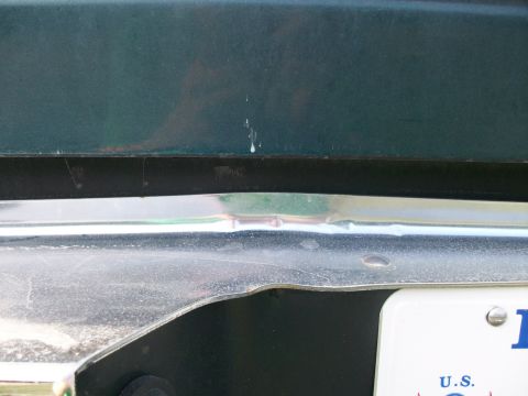 1995 Ford C-350 (Centurion Conversions) - Close-up of a dark green lower body panel meeting a dented and pitted chrome bumper, with rust bloom showing through the paint above the se…