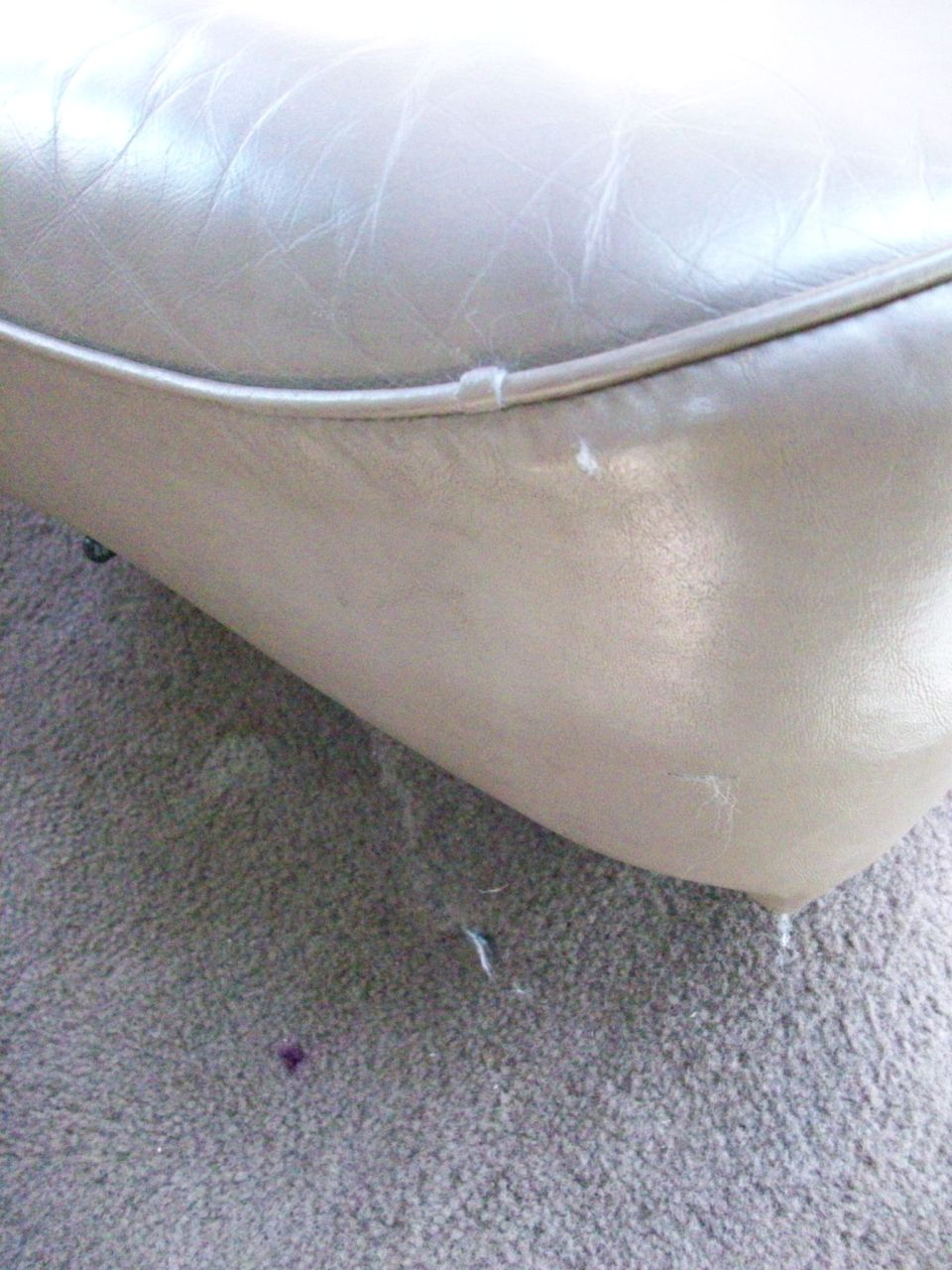 Close-up of a light tan, worn leather seat cushion with visible scuffs and loose threads. Carpet is visible below