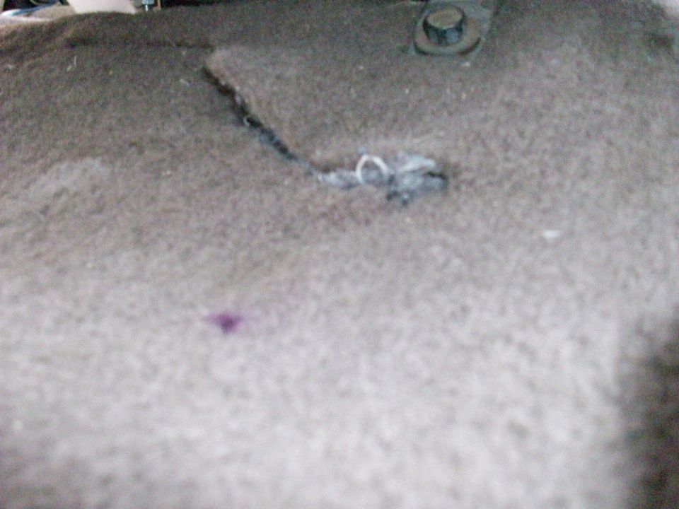 Torn and burned carpet on a vehicle floor near a bolt