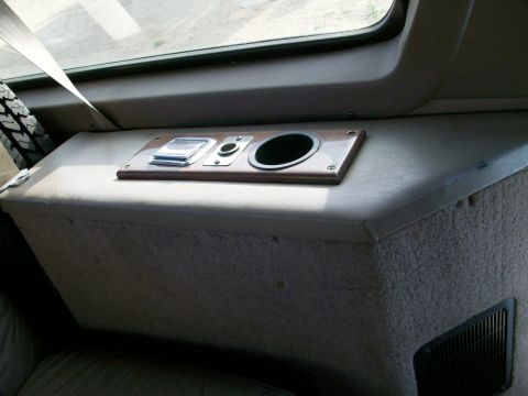 1995 Ford C-350 (Centurion Conversions) - Rear interior quarter panel of a conversion vehicle in tan vinyl and carpet, with a wood-grain trim plate holding an ashtray, a 12V power/l…