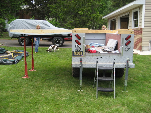 Homebuilt truck camper on jack stands beside aluminum flatbed utility body on lawn