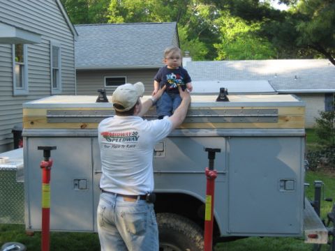 1996 Ford Bronco - A man lifts a baby onto the top of a utility truck bed, which is equipped with a wooden extension and metal hinges.