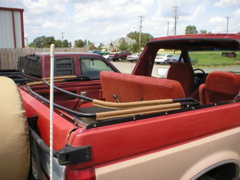 1983 Ford Bronco - This red convertible SUV is parked next to a maroon pickup truck under a partly cloudy sky.