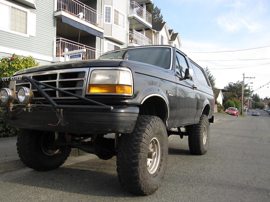 Black lifted Ford Bronco SUV with mud tires and grille guard, front 3/4 view on a street