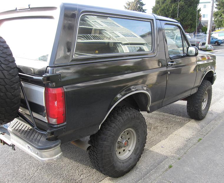 Black lifted Ford Bronco SUV with big off-road tires, rear passenger-side view at curb
