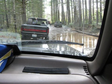1992 Ford Bronco - Multiple vehicles navigate a muddy path in a forest, with a 1992 Ford Bronco leading the way.