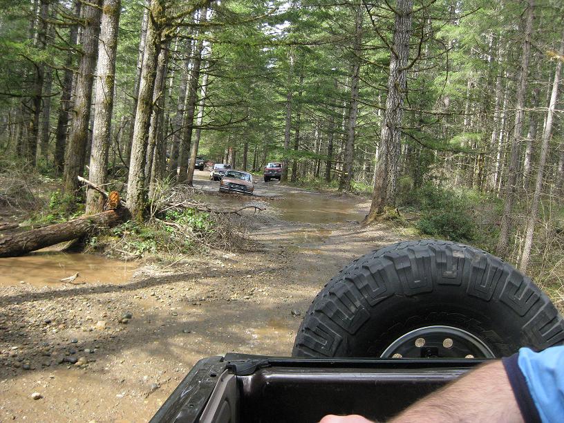 Several vehicles driving on a muddy forest road, with a spare tire visible in the foreground