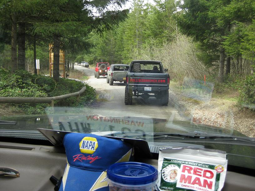 Several trucks driving on a dirt road through a forest. View from inside a vehicle