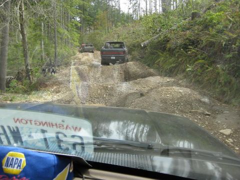 1992 Ford Bronco - Two trucks navigate a challenging dirt road through a wooded area, viewed from the perspective of another vehicle.