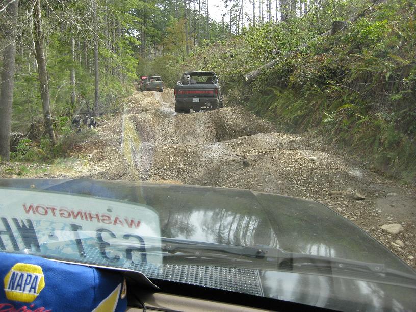 View from inside a vehicle looking at two trucks on a rough dirt road in a forest