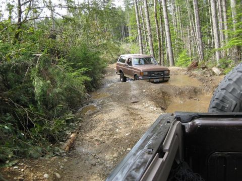 1992 Ford Bronco - A brown Ford Bronco tackles a challenging, muddy off-road trail surrounded by dense forest.