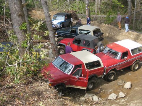 1992 Ford Bronco - Multiple off-road vehicles are seen on a wooded trail, with several people observing the action.
