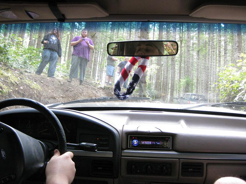 Interior view from driver's seat of a vehicle on a forest road; people stand on the embankment. Another vehicle is visible in the distance