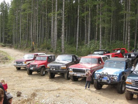 1992 Ford Bronco - A collection of off-road vehicles are gathered on a dirt path surrounded by tall trees.