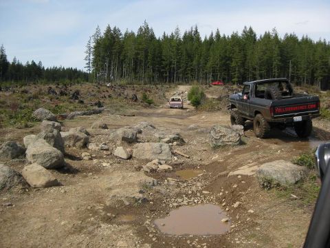 1992 Ford Bronco - A black Ford Bronco navigates a rocky, wooded off-road course, with another vehicle visible in the distance.