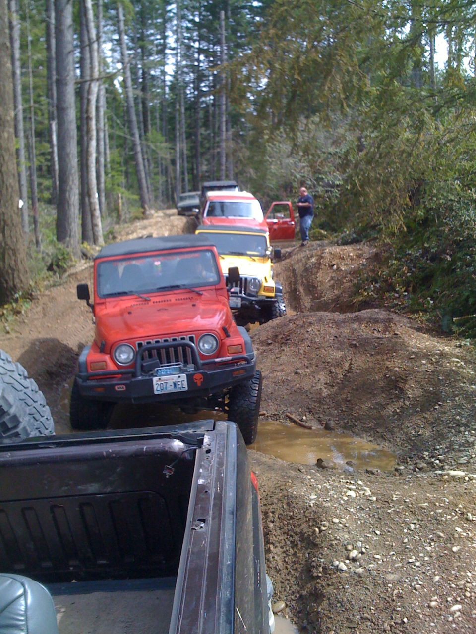Red Jeep Wrangler driving on a muddy forest trail with other vehicles behind it
