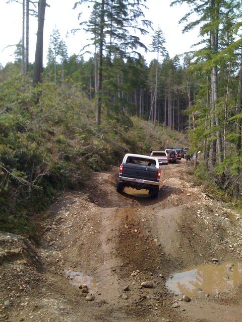 1992 Ford Bronco - A convoy of Ford Broncos navigates a challenging, muddy trail through a dense forest.