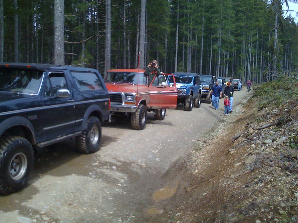 Several off-road vehicles driving down a dirt road in a forest. A red truck has its door open
