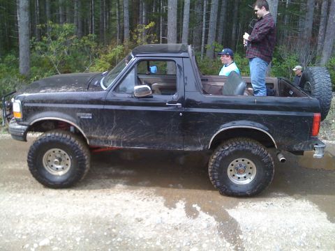 1992 Ford Bronco - A 1992 Ford Bronco with a lifted suspension and off-road tires is seen on a dirt path with trees in the background.