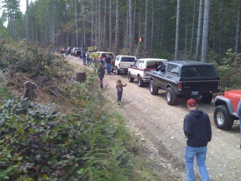 1992 Ford Bronco - Several off-road vehicles are lined up on a dirt path surrounded by tall trees.