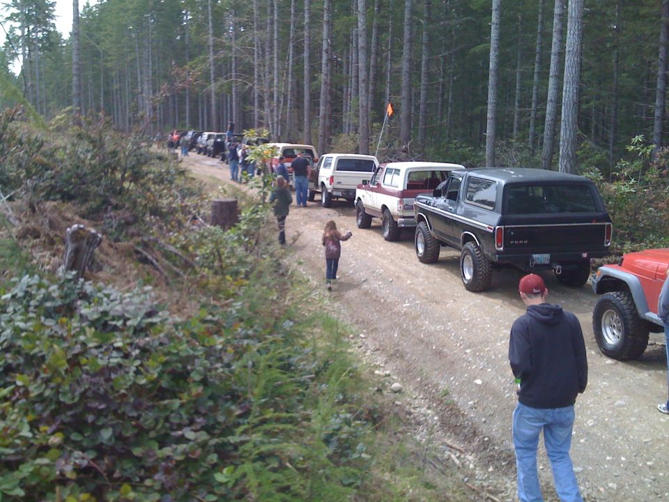 Line of SUVs and Jeeps parked on a dirt road in a forest. A black Ford Bronco is in the foreground