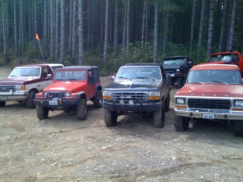 1992 Ford Bronco - Several trucks and SUVs are parked in a dirt lot with a forest in the background.