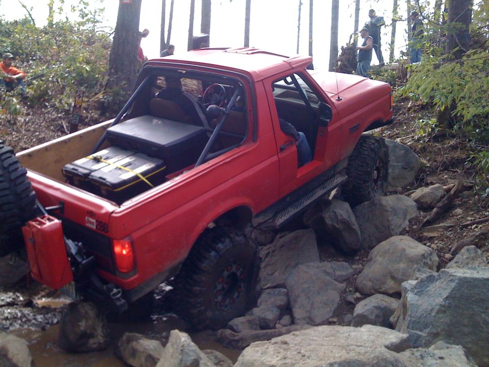 Red 1992 Ford Bronco off-roading over rocks, with a roll cage and extra fuel cans