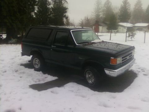 1990 Ford Bronco - A dark green Ford Bronco sits in a snowy yard, its body style indicating a model year around 1987.
