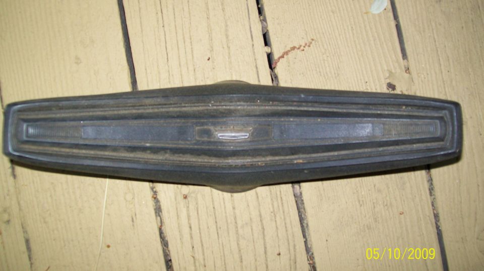 Dark gray car emblem and trim piece with horizontal lines and a central badge, resting on wooden planks