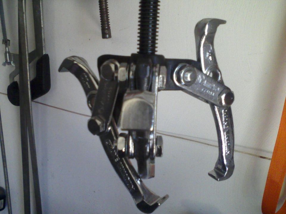 Gear puller tool with three jaws and a threaded shaft, hanging against a wall