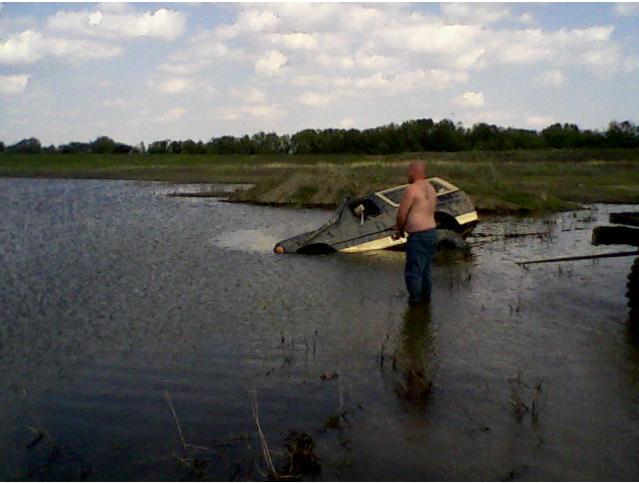 Man stands in shallow water next to a partially submerged SUV, which appears to be stuck