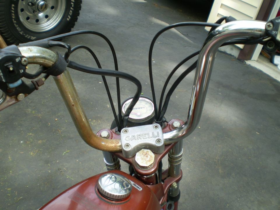 Close-up of Garelli moped handlebars, fuel cap, and speedometer. A tire is visible in the background