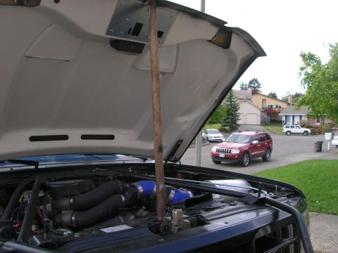 1996 Ford Bronco - The engine bay of a dark vehicle is visible with the hood propped open by a wooden stick, showing blue hoses and other components.