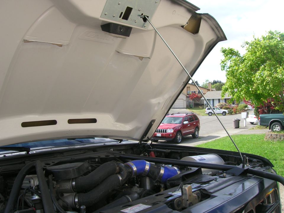 Open tan hood over an engine bay with blue silicone intake couplers, red Jeep parked across the street