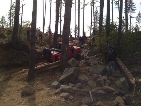 1989 Ford Bronco - A red off-road vehicle is navigating a difficult rocky trail in a wooded area, with several people observing the action.