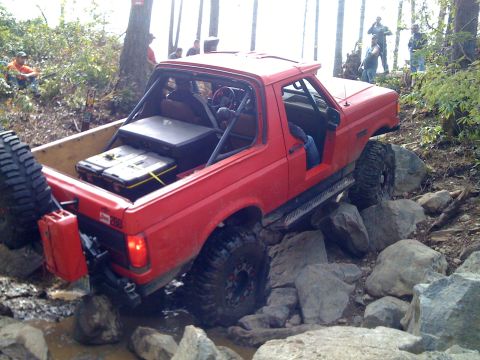 1989 Ford Bronco - This modified SUV is shown off-roading through a rocky, wooded area with several people watching.
