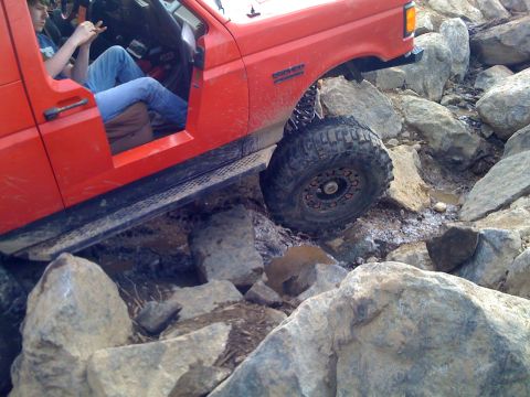 1989 Ford Bronco - This red SUV is shown off-roading over a rocky terrain, with one wheel elevated.