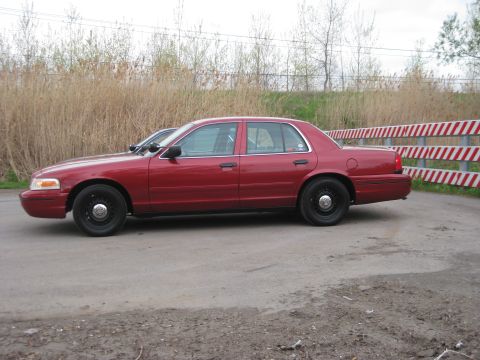 2000 Ford Crown Victoria - Side view of a red sedan parked on a dirt road with a striped barrier in the background.
