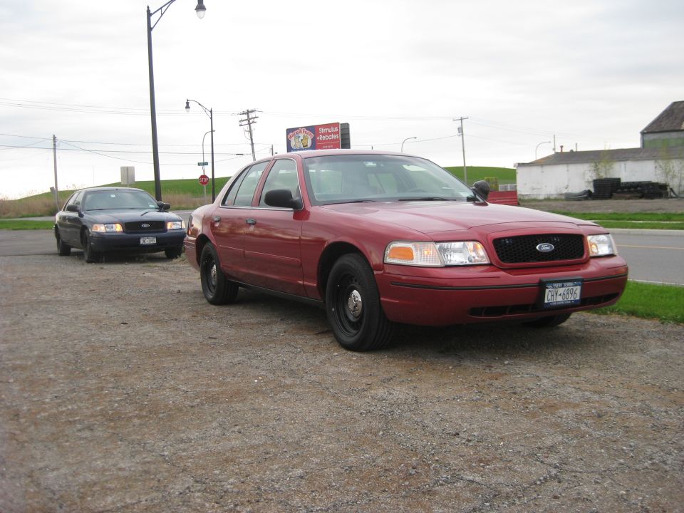 Red Ford Crown Victoria, front 3/4 view, parked on gravel lot with another dark Crown Victoria behind it