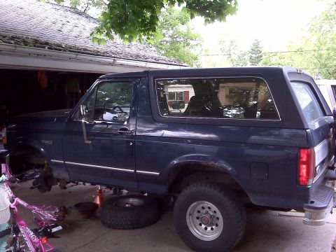 1995 Ford Bronco - A dark blue Ford Bronco is parked in a driveway with its front end disassembled and raised on jacks.