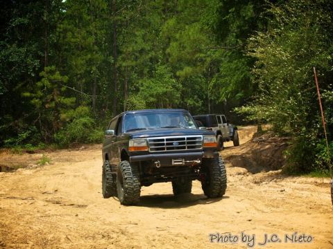 1995 Ford Bronco - Two off-road vehicles, a Ford Bronco and a Jeep Wrangler, navigate a sandy trail through a wooded area.