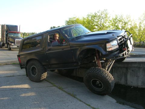 1996 Ford Bronco - A modified black 1996 Ford Bronco is shown with its front tire on a concrete wall, while a semi-truck is visible in the background.
