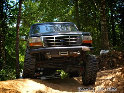 1995 Ford Bronco - A lifted black Ford Bronco navigates an off-road trail surrounded by trees.