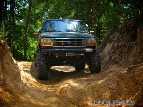 1995 Ford Bronco - This lifted Ford Bronco navigates a challenging off-road trail surrounded by trees.
